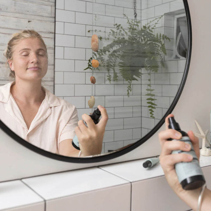 Person applying mist in front of a mirror with a plant in the background.