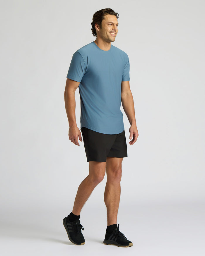 A man wearing a light blue short-sleeve athletic t-shirt, black athletic shorts, and black sneakers, standing against a light gray studio background.