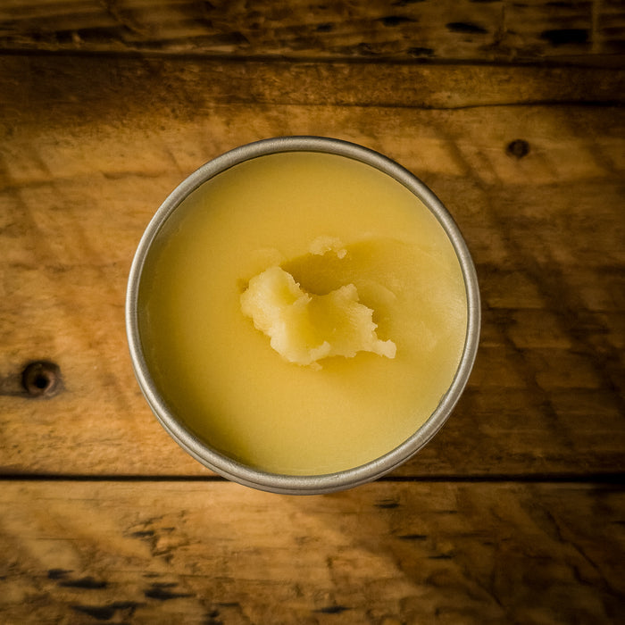 Top view of an open tin of Mammoth Beard Co. beard balm on a rustic wooden surface, highlighting its smooth, natural formula with a small portion scooped out.