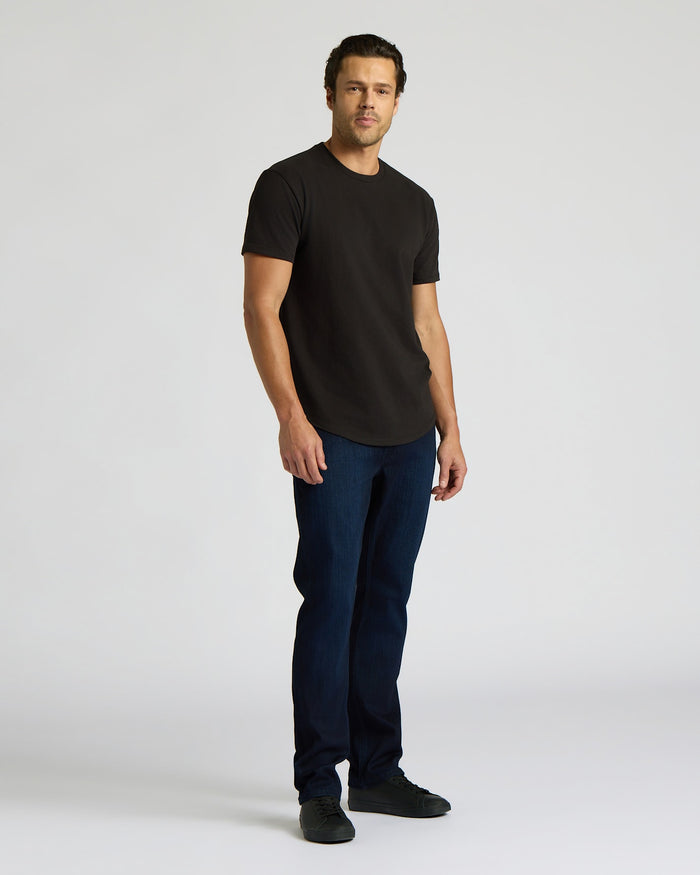 Man wearing a black Heavyweight Curved Hem Crew 6-Pack t-shirt paired with dark blue jeans and black shoes, standing against a plain light background.