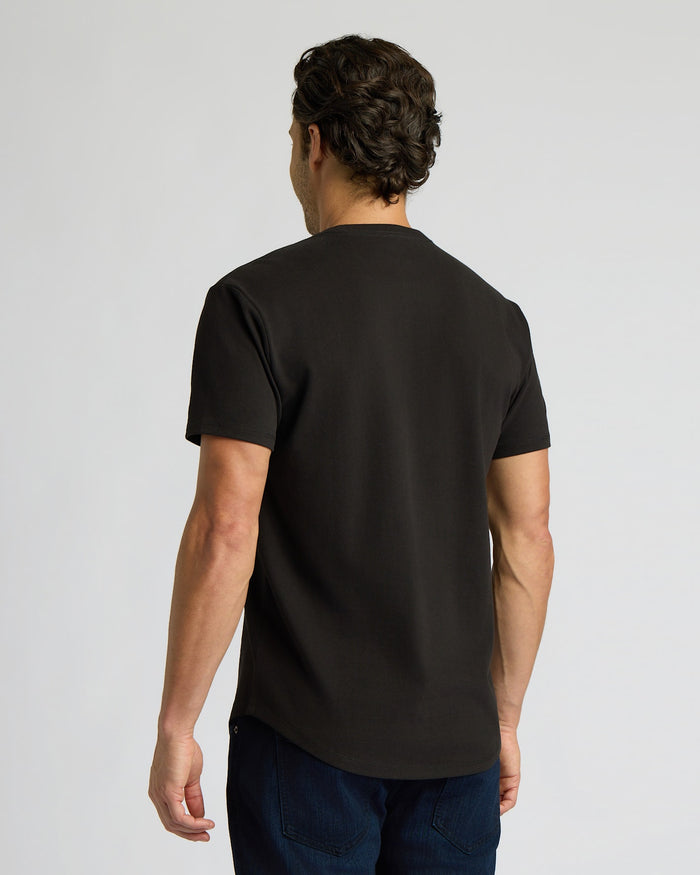 Man wearing an All Black Heavyweight Curved Hem Crew 6-Pack t-shirt, viewed from the back, showcasing the curved hem and heavyweight fabric in a simple studio setting.