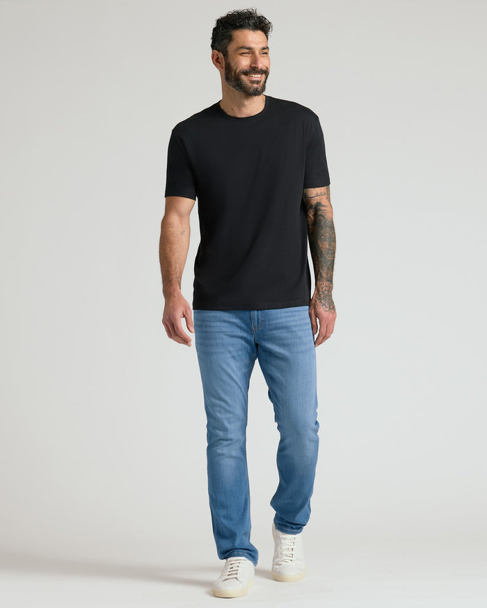 Man wearing a black Pima Cotton Blend Crew 4-Pack t-shirt paired with light blue jeans and white sneakers, smiling and walking confidently against a plain background