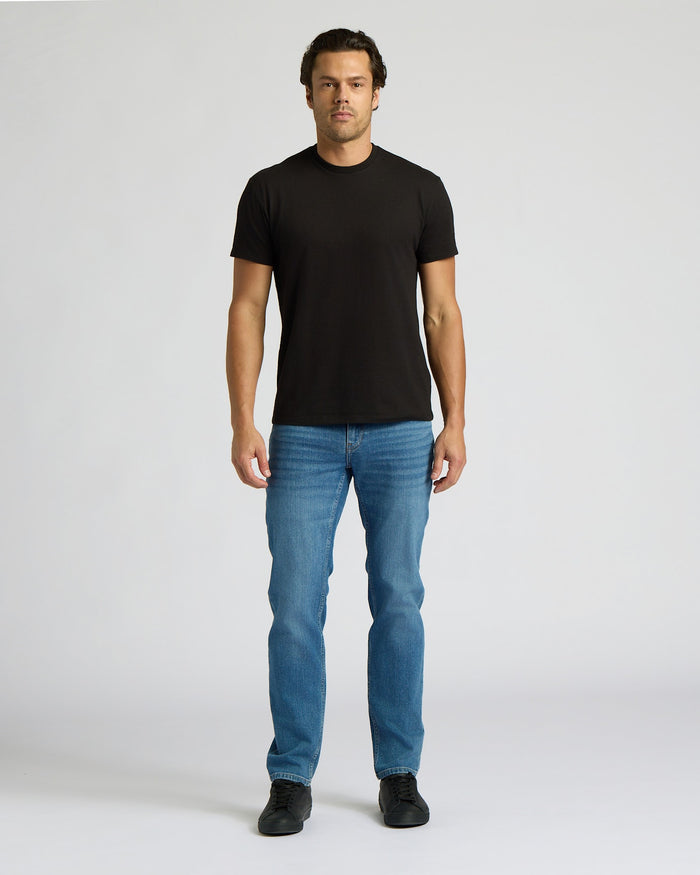Man wearing a black t-shirt and blue jeans on a white background. MODEL_INFO: Model is 6'0" wearing size L short