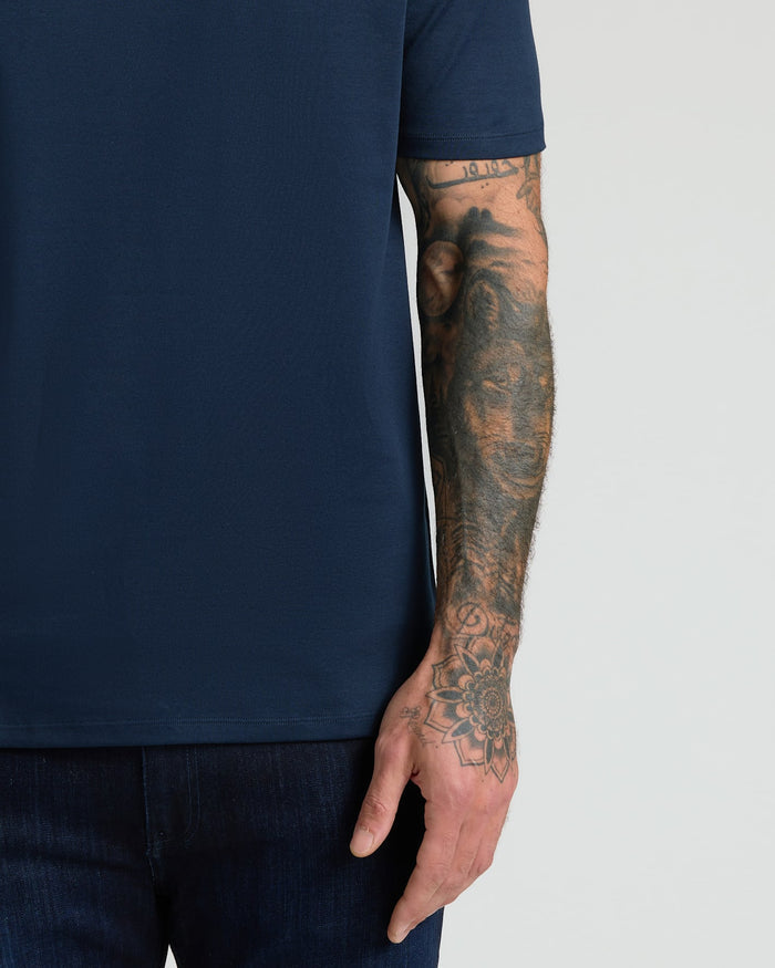 Close-up of a man's arm with detailed tattoos wearing a Navy Pima Cotton Blend Crew shirt paired with dark jeans against a plain light background.