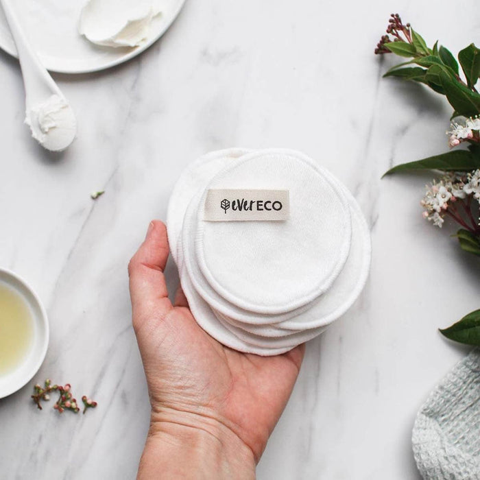 Hand holding a pile of bamboo facial pads with skincare products on a marble surface with flowers.