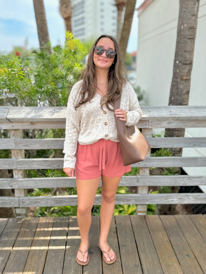 Woman in a white cardigan, pink shorts and sunglasses standing on a wooden deck.