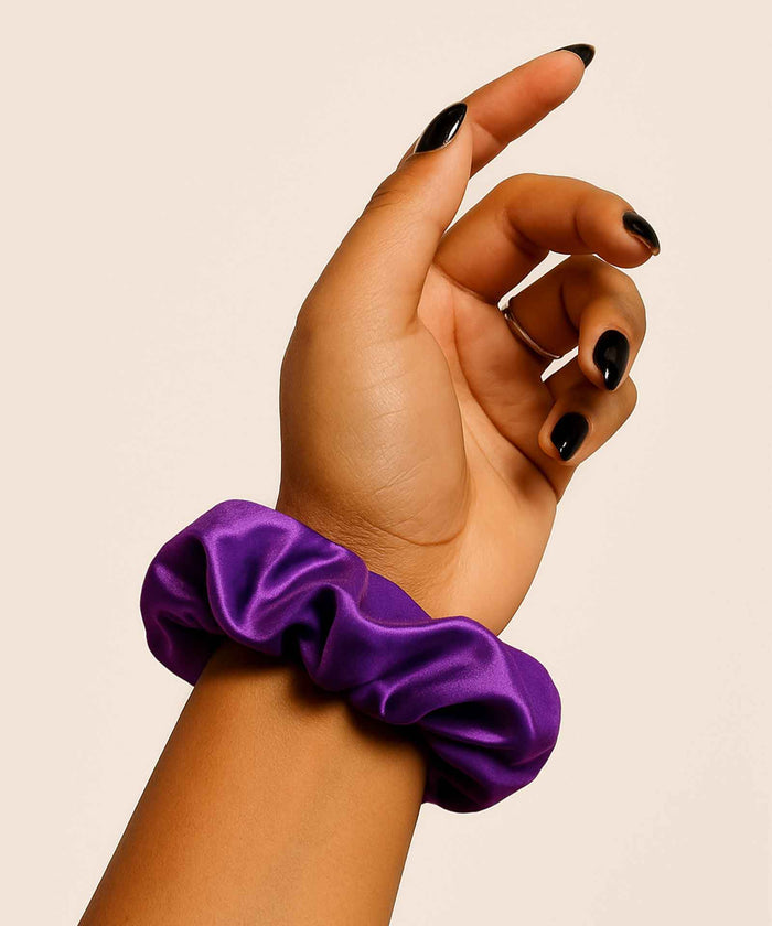 A hand with black-painted nails is raised against a light background, adorned with a shiny purple velvet scrunchie around the wrist.