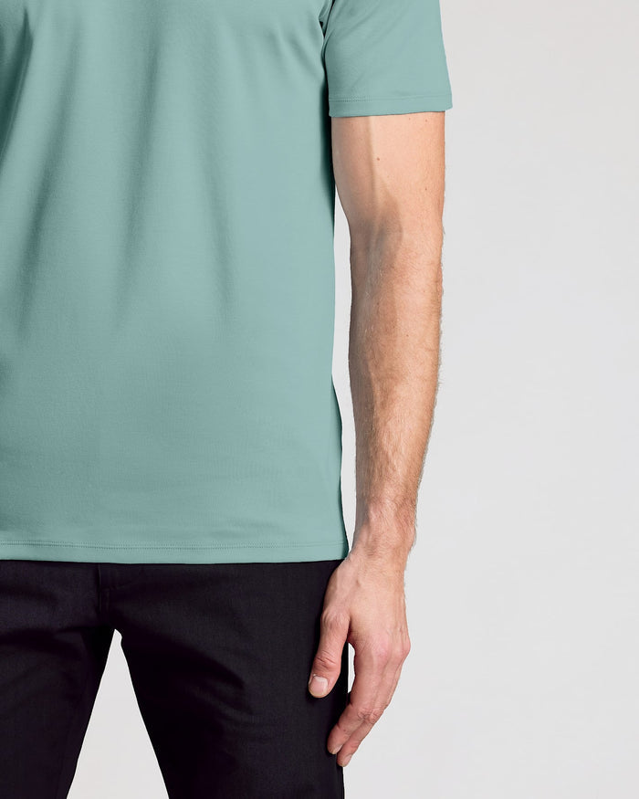 A close-up of a man wearing a short-sleeve crewneck t-shirt in a muted sage green color, paired with black pants, against a light gray background.