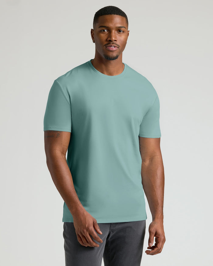 A man wearing a solid sage green crew-neck t-shirt and dark gray pants, standing against a light gray studio background.