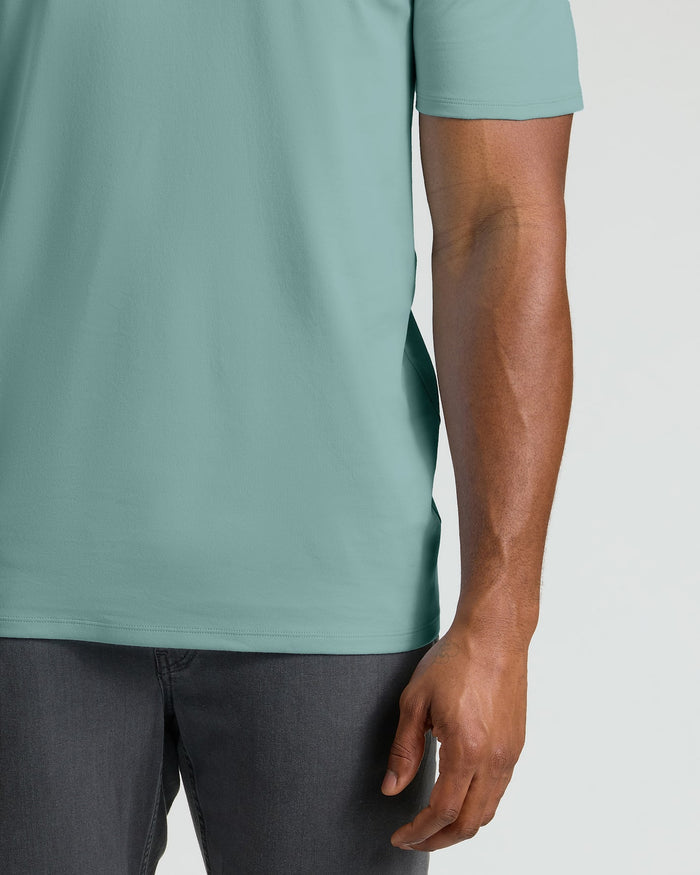 A close-up of a person wearing a short-sleeve crew-neck t-shirt in a muted sage green color, paired with dark gray jeans, against a light gray background.
