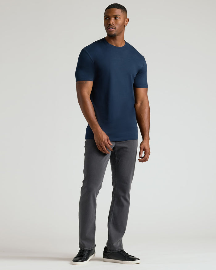 A man wearing a navy blue Tall Pima Cotton Blend Crew 4-Pack t-shirt paired with gray pants and black sneakers, standing against a plain light background.
