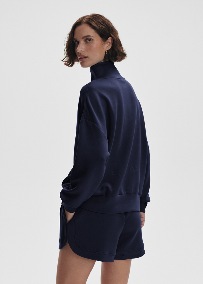 Woman turned to the side wearing a navy half zip sweatshirt top with matching shorts in front of a white background
