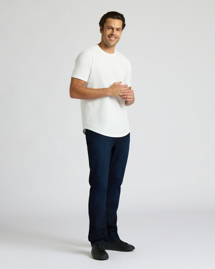 Man wearing a white Heavyweight Curved Hem Crew 6-Pack t-shirt paired with dark jeans and black shoes, standing and smiling against a plain background.