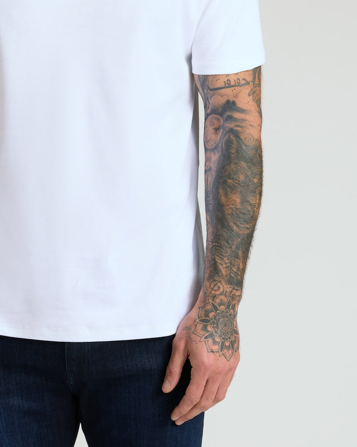 Close-up of a man's arm with intricate tattoos wearing a white short-sleeve shirt from the Pima Cotton Blend Crew 2-Pack paired with dark jeans, showcasing men's basics.