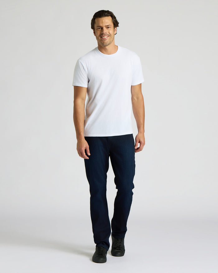 Man wearing a white t-shirt and dark jeans on a plain background. MODEL_INFO: Model is 6'0" wearing size L short