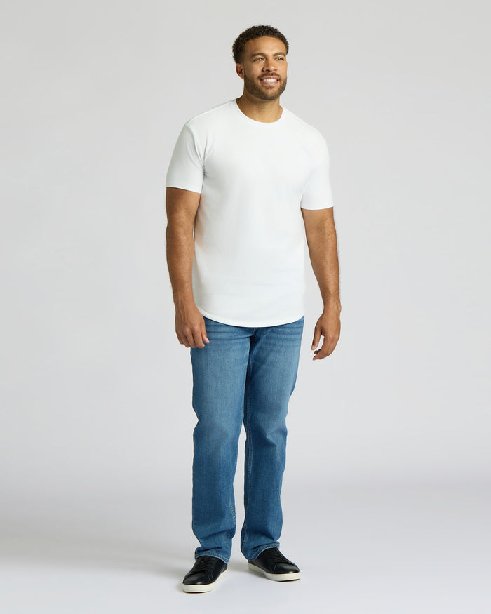 Man wearing an All White Tall Heavyweight Curved Hem Crew 3-Pack T-shirt paired with blue jeans and black shoes, standing against a light gray background.